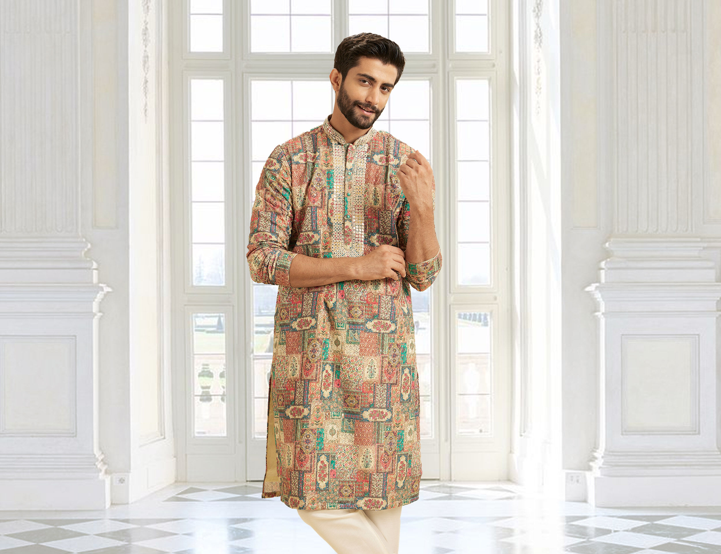 Man wearing a multi-colored printed kurta pajama set, standing in a grand hall with classic white interiors and tall French windows.